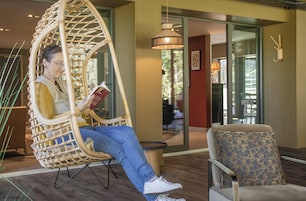 Una persona sentada en una silla de mimbre leyendo un libro en una habitación con decoración moderna, una mesita redonda y un sillón estampado.