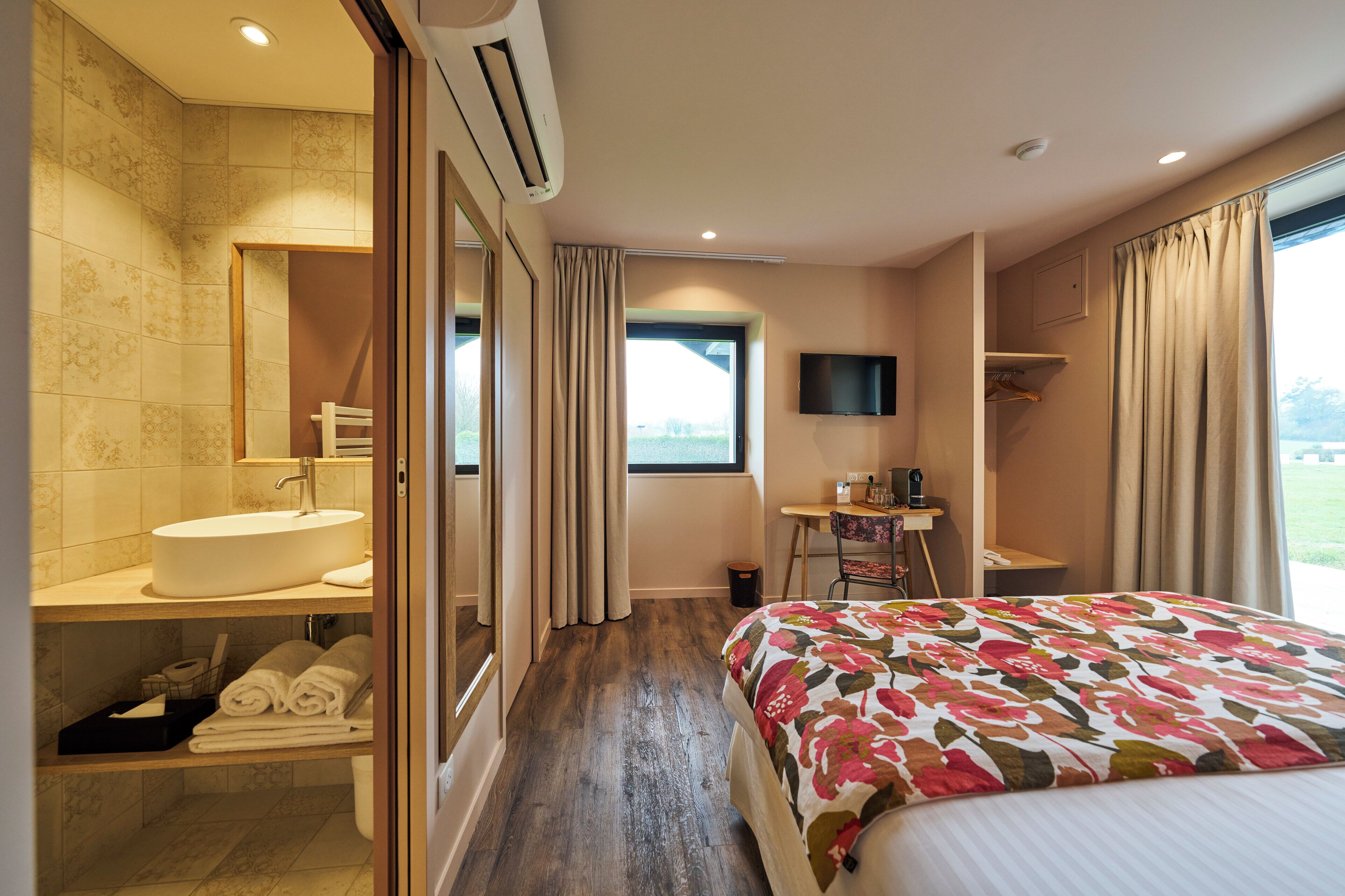 A hotel room with a large bed, a bathroom with a sink and mirror, a TV, and a window with curtains.