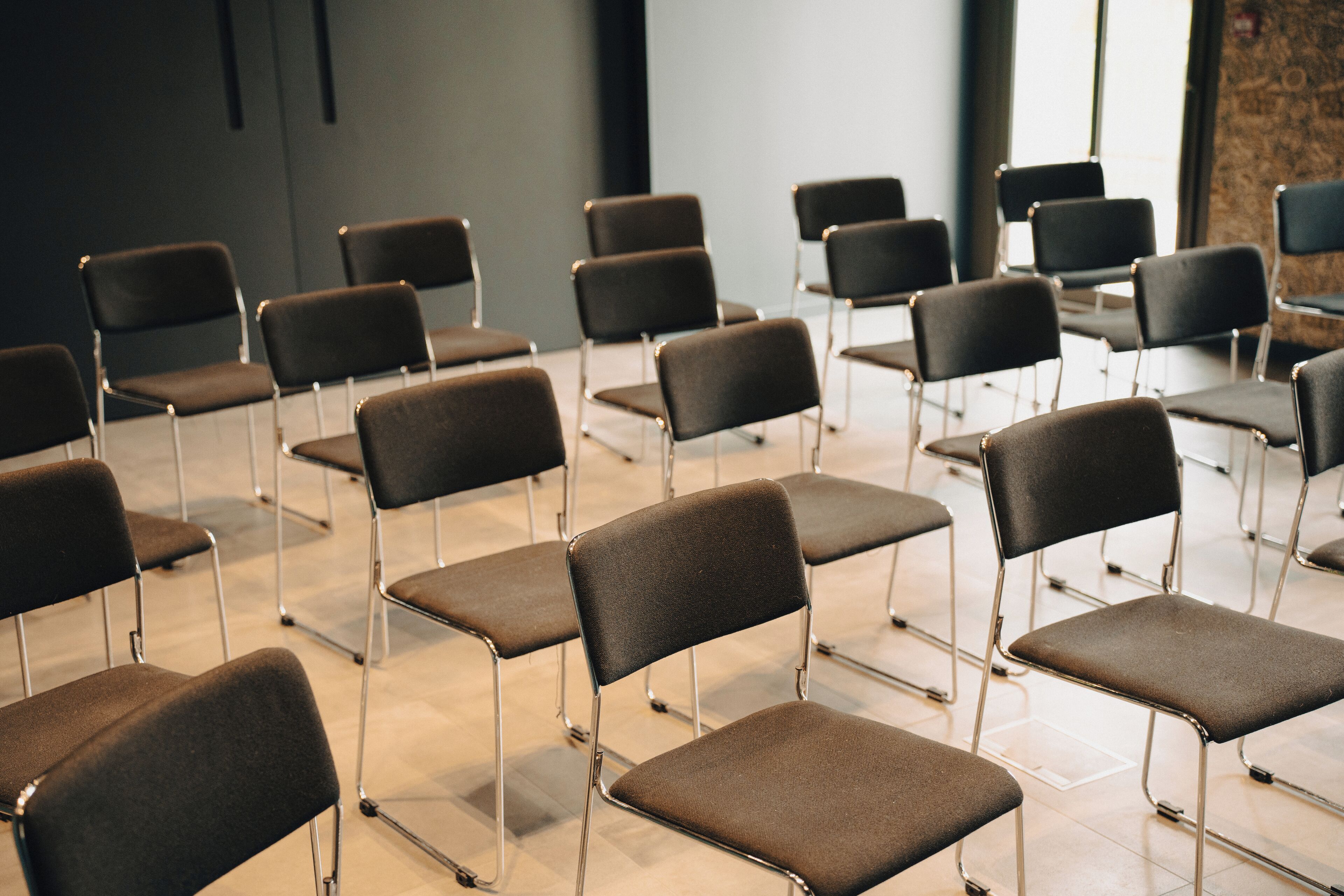 Des rangées de chaises sont disposées dans une salle de conférence, avec un mur sobre et une fenêtre en arrière-plan.