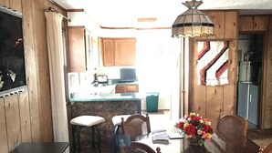 A dining area with a wooden table and chairs, a chandelier, and a kitchen in the background.