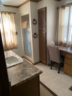 Un baño con lavamanos, ducha e inodoro. Hay una silla y un escritorio con una computadora.