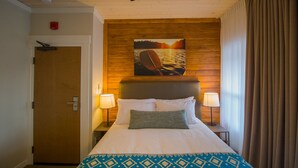 A hotel room with a wooden headboard, a painting of a sunset over water, two bedside lamps, and a patterned bedspread. - Cranberry Arms Inn (Cassidy)