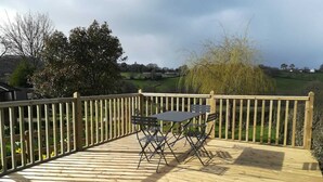 Een houten terras met een tafel en stoelen, omgeven door een houten hek, met uitzicht op een landelijk gebied.