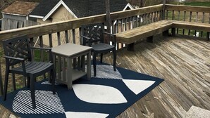 Una terraza de madera con una mesa y sillas, rodeada de árboles y un cobertizo pequeño.