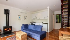 A living room with a sofa, a fireplace, a kitchen area, and a bookshelf. - Cloudsong Chalet 1 - Close to the village centre! (Kangaroo Valley)