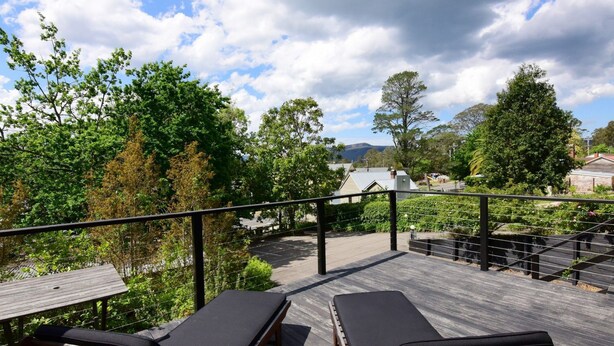 A wooden deck with lounge chairs, surrounded by lush greenery and a view of buildings and mountains in the distance. - Cloudsong Chalet 1 - Close to the village centre! (Kangaroo Valley)