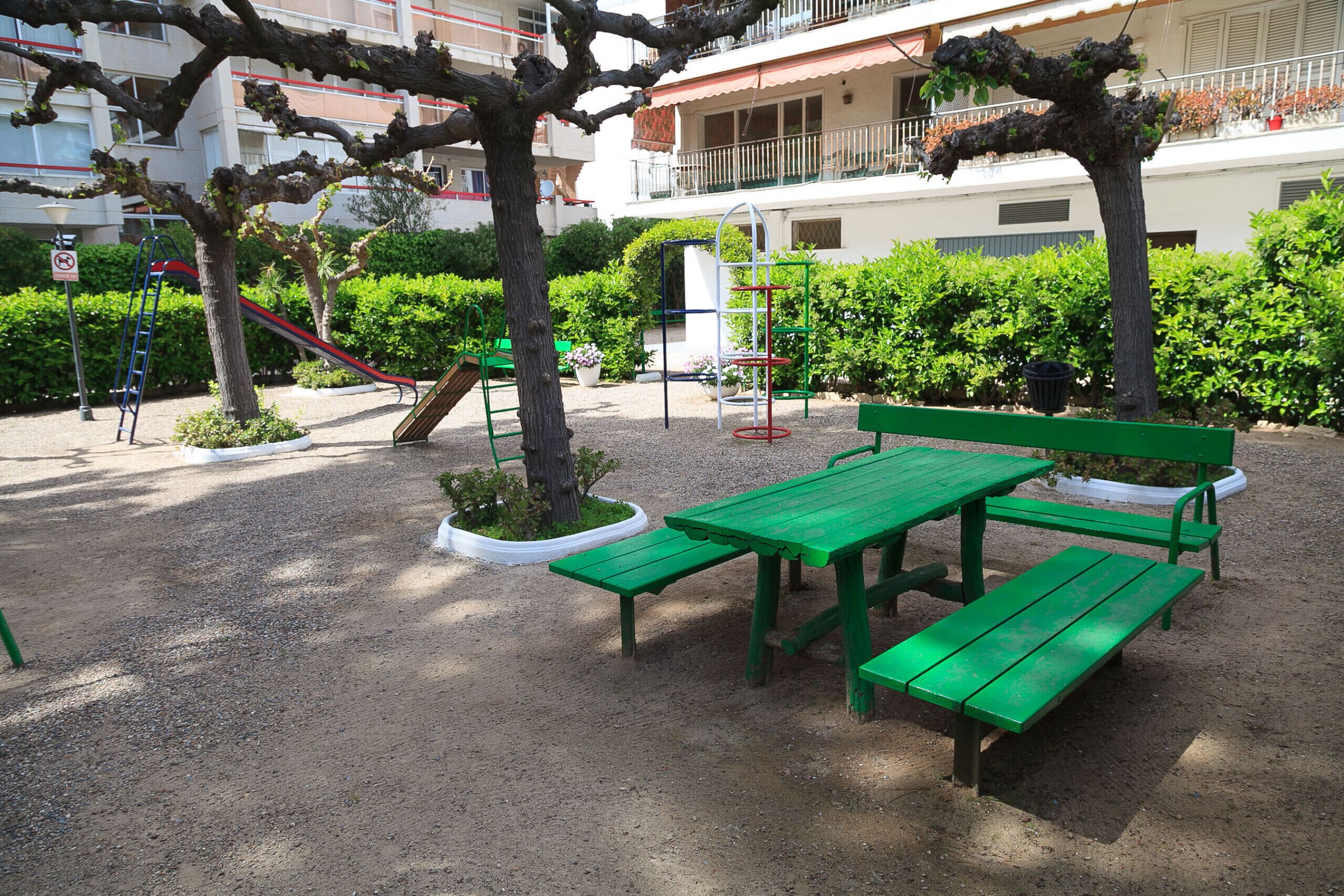 Un parque infantil con bancos verdes y un tobogán, rodeado de árboles y un edificio al fondo.