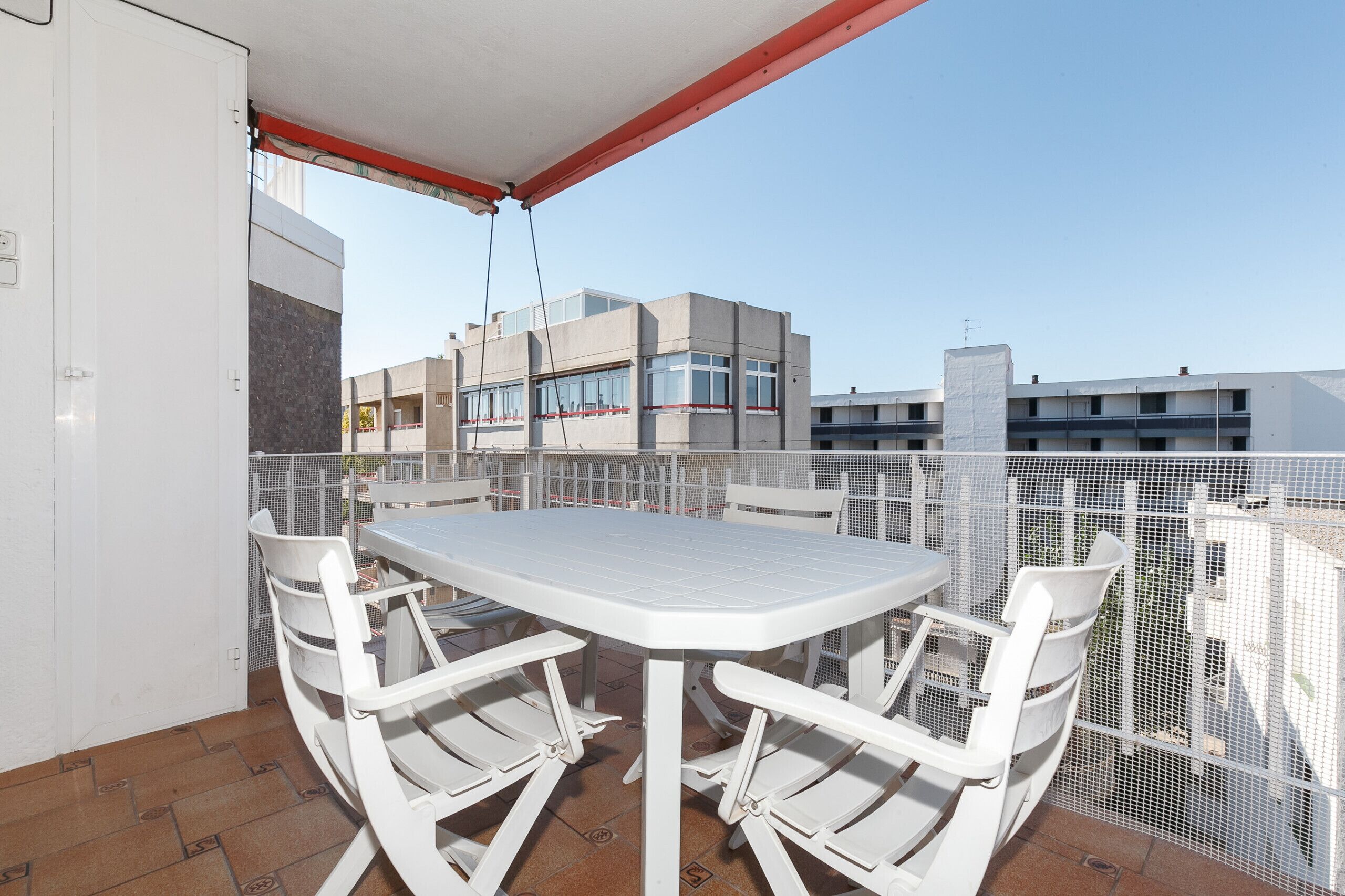 En balkon med et hvidt bord og stole, en rød markise samt udsigt til bygninger i baggrunden.