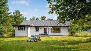 A small, single-story house with a tiled roof, a front porch with a table and chairs, and a lawn with two lounge chairs. - Awesome home in Knebel with kitchen (Knebel)