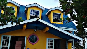 A multi-story building with a yellow and blue exterior, a central circular window, and a staircase leading to the entrance. - Tropickeas (St. George's Caye)
