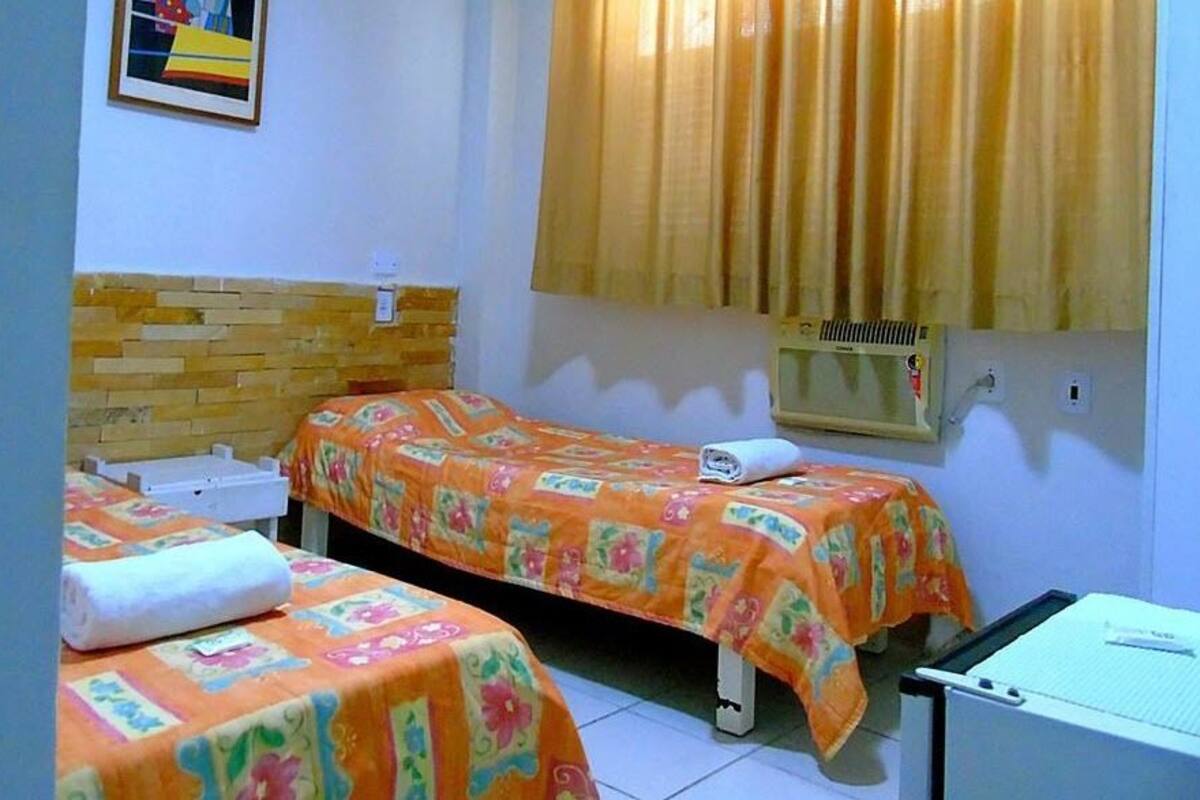 Ein kleines Zimmer mit zwei Betten, jeweils mit farbenfroher Blumendecke und weißem Kopfkissen. Ein kleiner Tisch mit weißem Tuch und ein Heizkörper an der Wand.