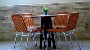 A dining area with a table and four wicker chairs, a potted plant on the table, and a stone wall in the background. - Pousada Xamã Tour (João Pessoa)