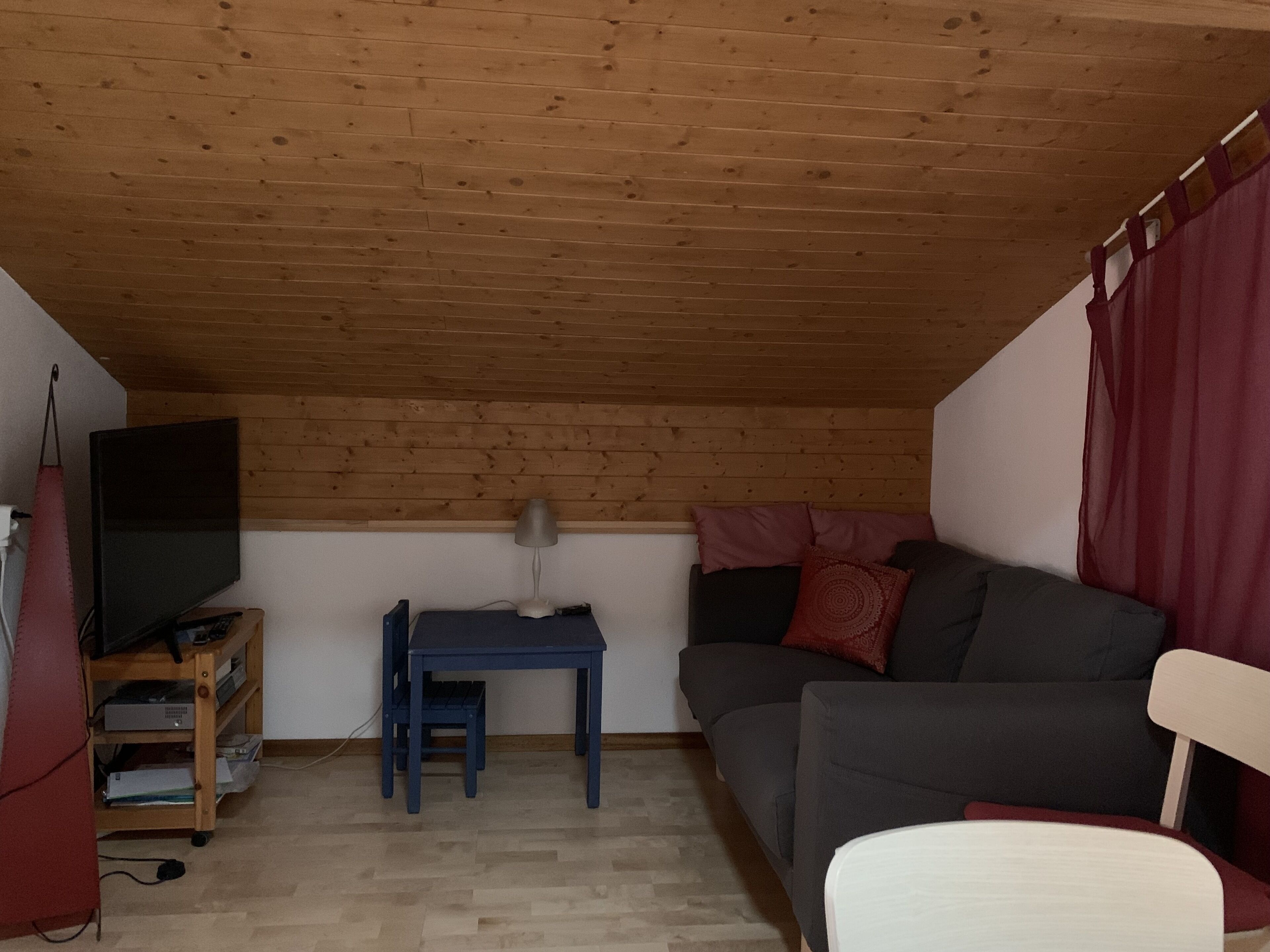 Ein gemütliches Schlafzimmer im Loft-Stil mit Holzbalkendecke, einem grauen Sofa, einem kleinen Tisch, einem Fernseher und einem roten Vorhang.