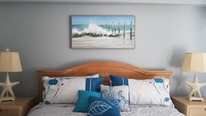 A bedroom with a wooden headboard, floral pillows, a starfish nightstand lamp, and a painting of a beach scene.