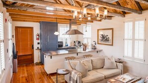 A cozy living room with a wooden ceiling, a sofa, a coffee table, and a kitchen area in the background. - King Bed Suite | Claw-Foot Tub, Chef Kitchen & Parking | Downtown Charleston (Charleston)