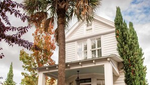 A two-story house with a porch, a palm tree, and a tall evergreen tree. - King Bed Suite | Claw-Foot Tub, Chef Kitchen & Parking | Downtown Charleston (Charleston)