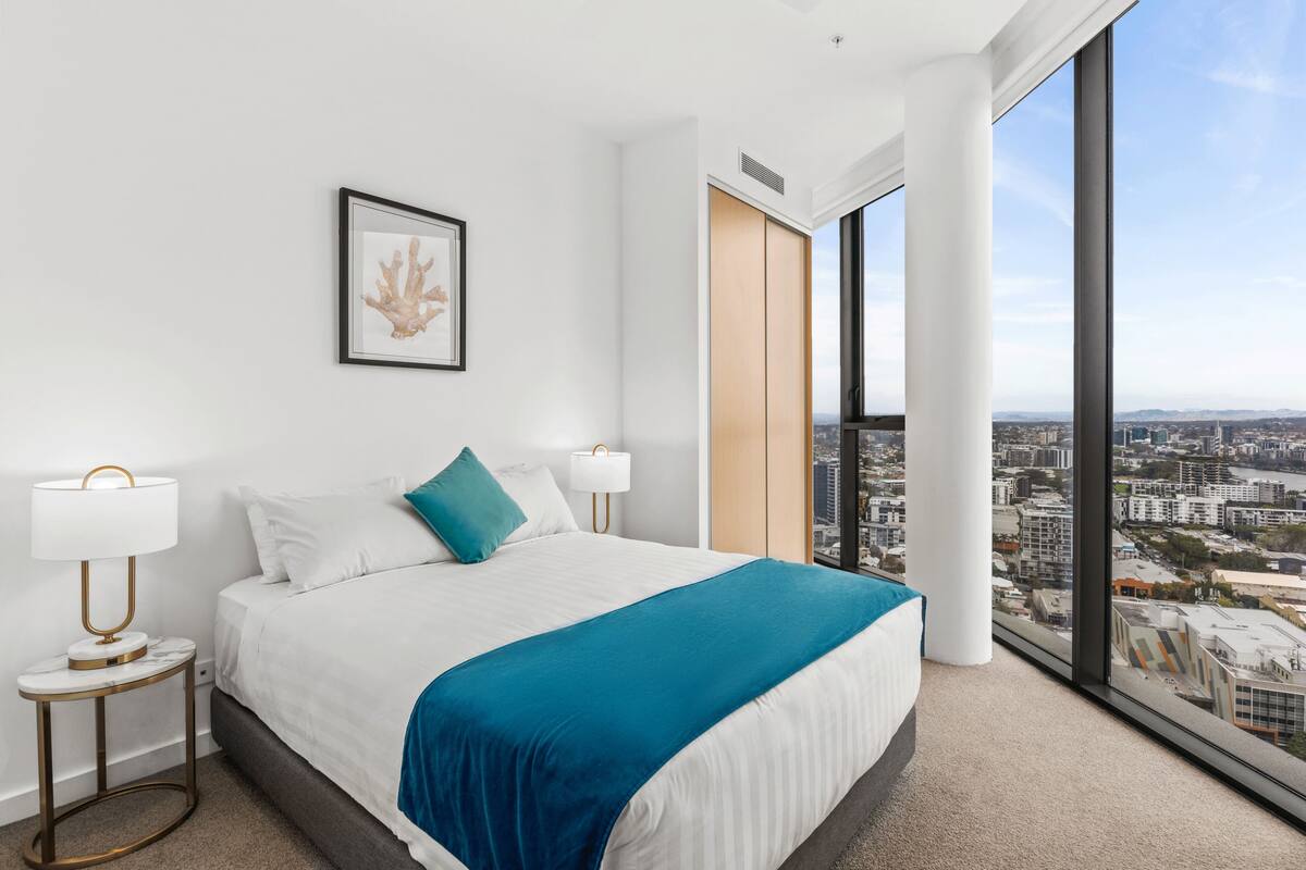 Una habitación de hotel moderna con una cama grande, dos mesitas de noche con lámparas, una obra de arte enmarcada en la pared y vistas a la ciudad a través de amplios ventanales.