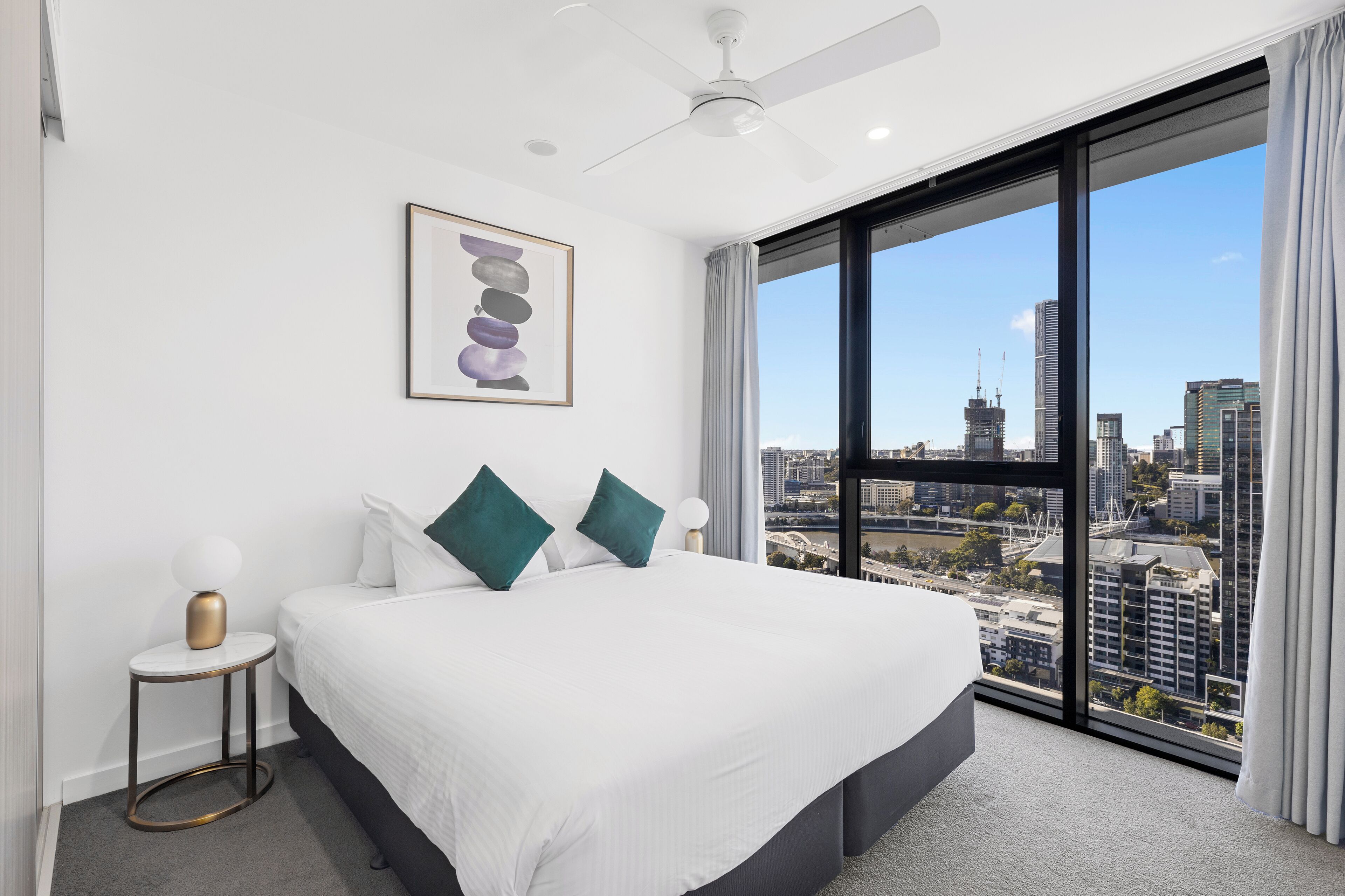 A modern hotel room with a large bed, a city view, and a ceiling fan.