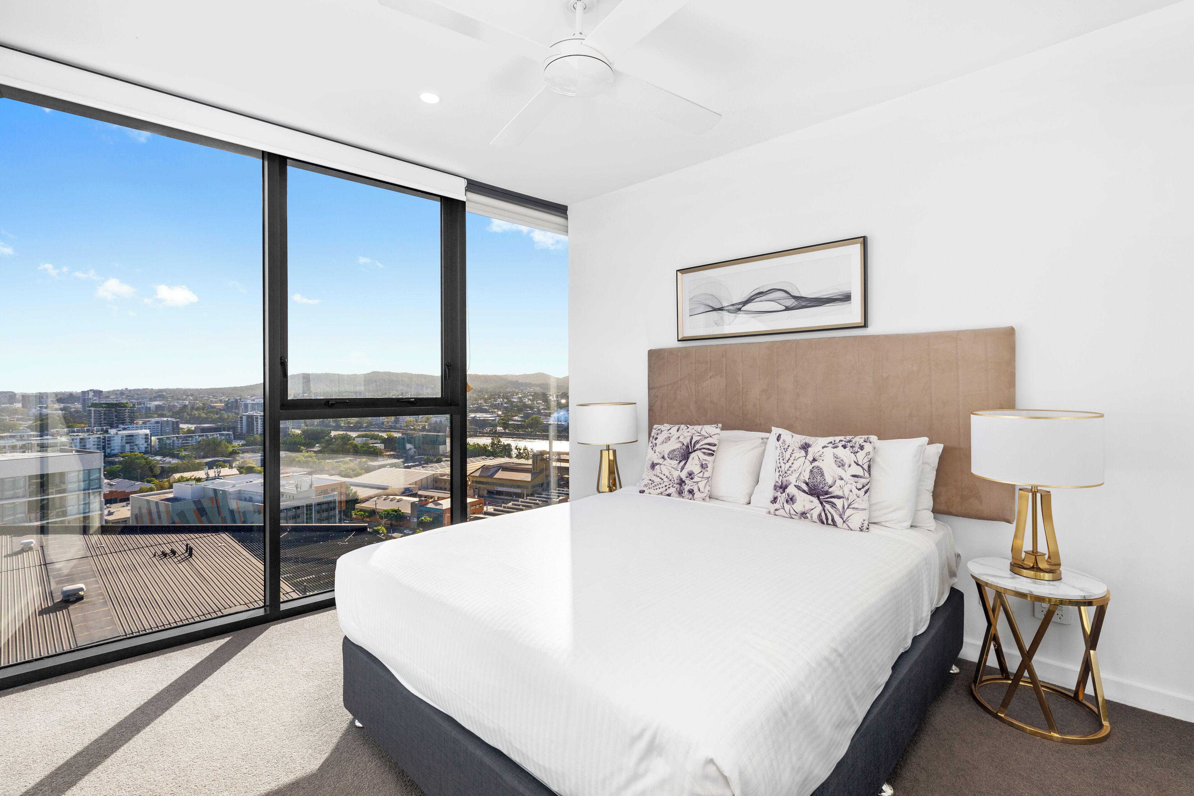 A modern hotel room with a large bed, a city view through floor-to-ceiling windows, a ceiling fan, and a framed artwork above the bed.