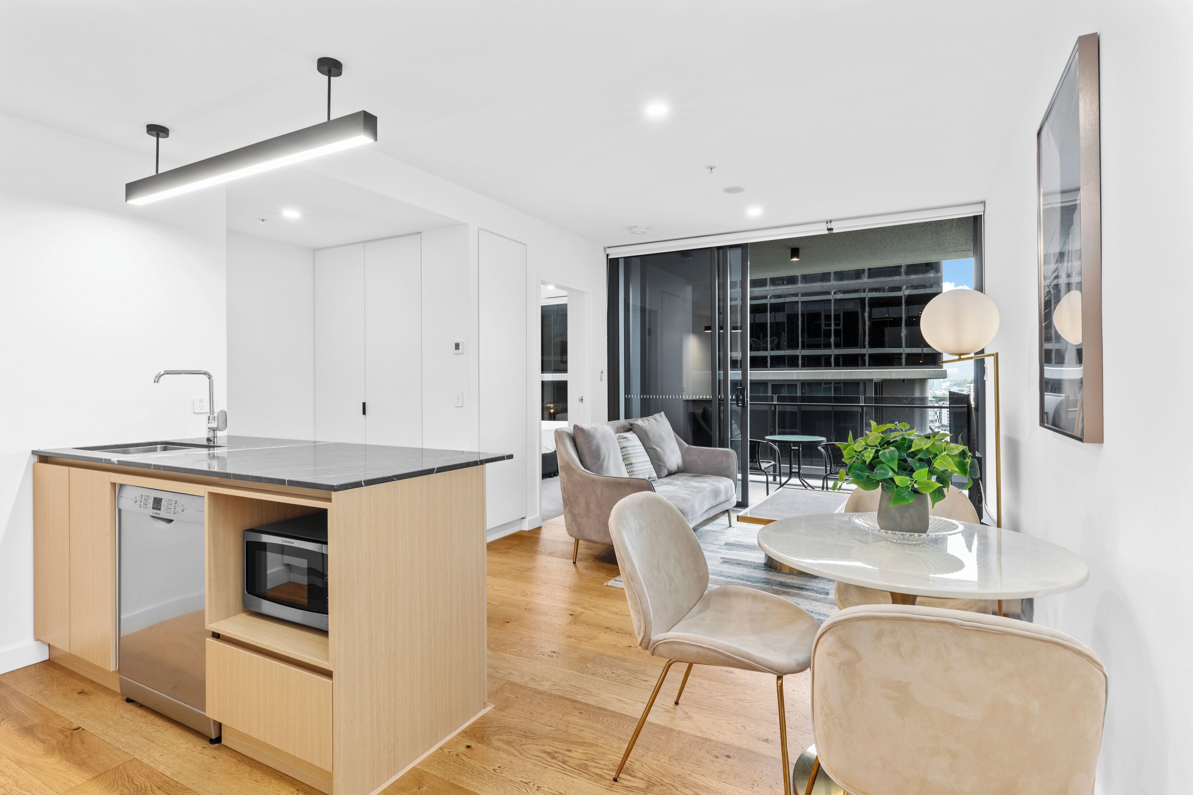 A modern kitchen with a dishwasher, microwave, and a central island. There is a dining area with a table and chairs, and a large window offering a view of the city.