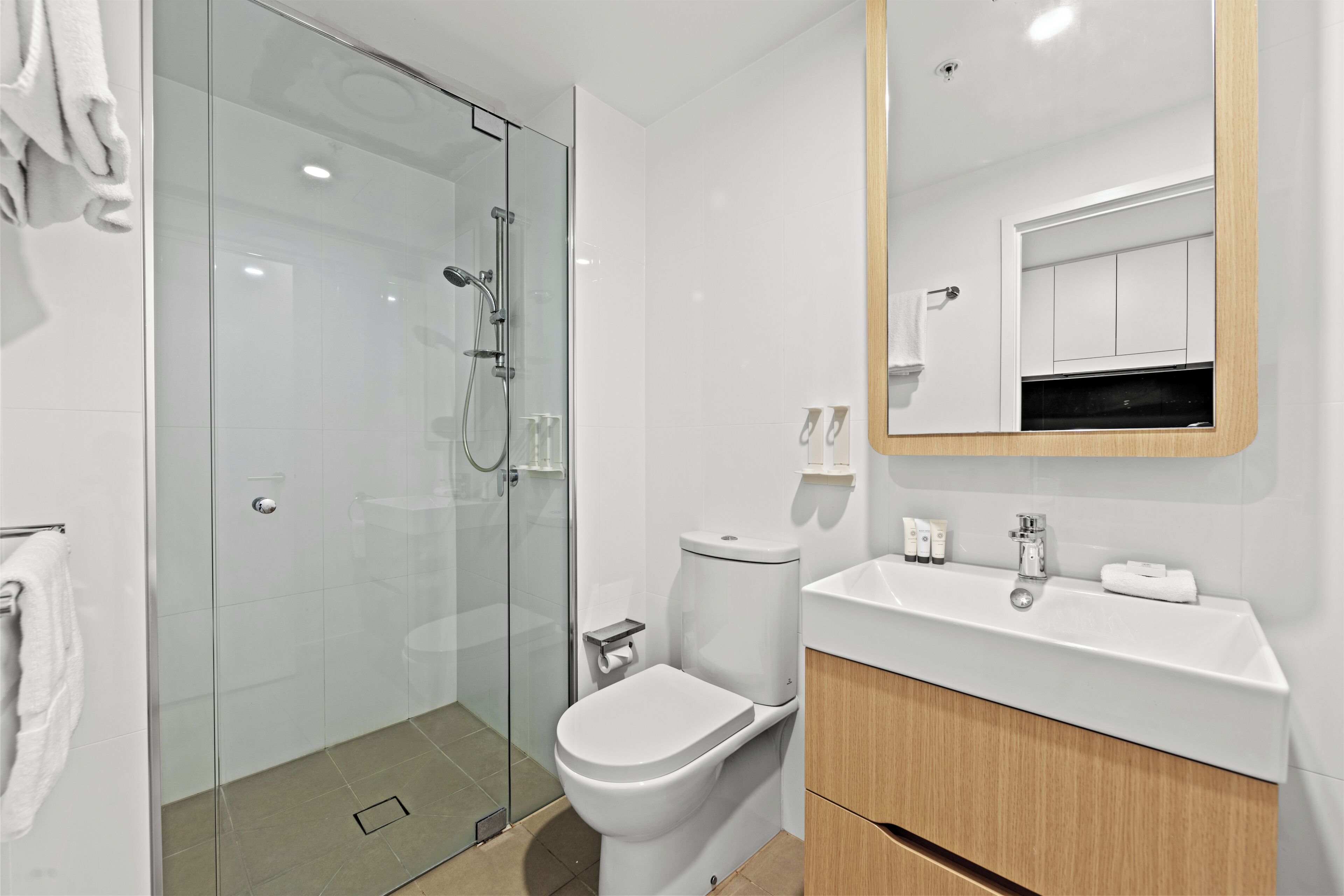 Un baño moderno con cabina de ducha de vidrio, inodoro blanco, lavabo con espejo y tocador de madera.