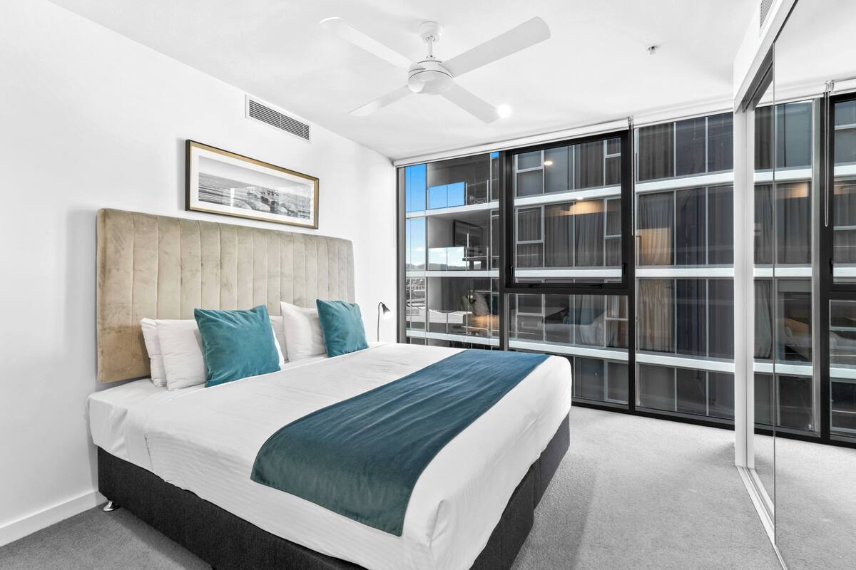 Un dormitorio moderno con una cama grande, un ventilador de techo y vistas a un edificio a través de ventanales de pared a pared.