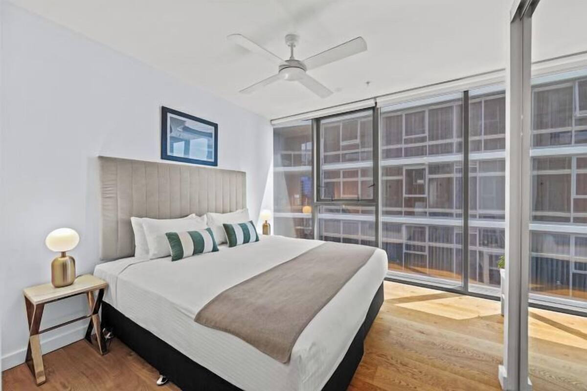 Un dormitorio con cama, mesita de noche, ventilador de techo y una ventana grande con vista a un edificio.