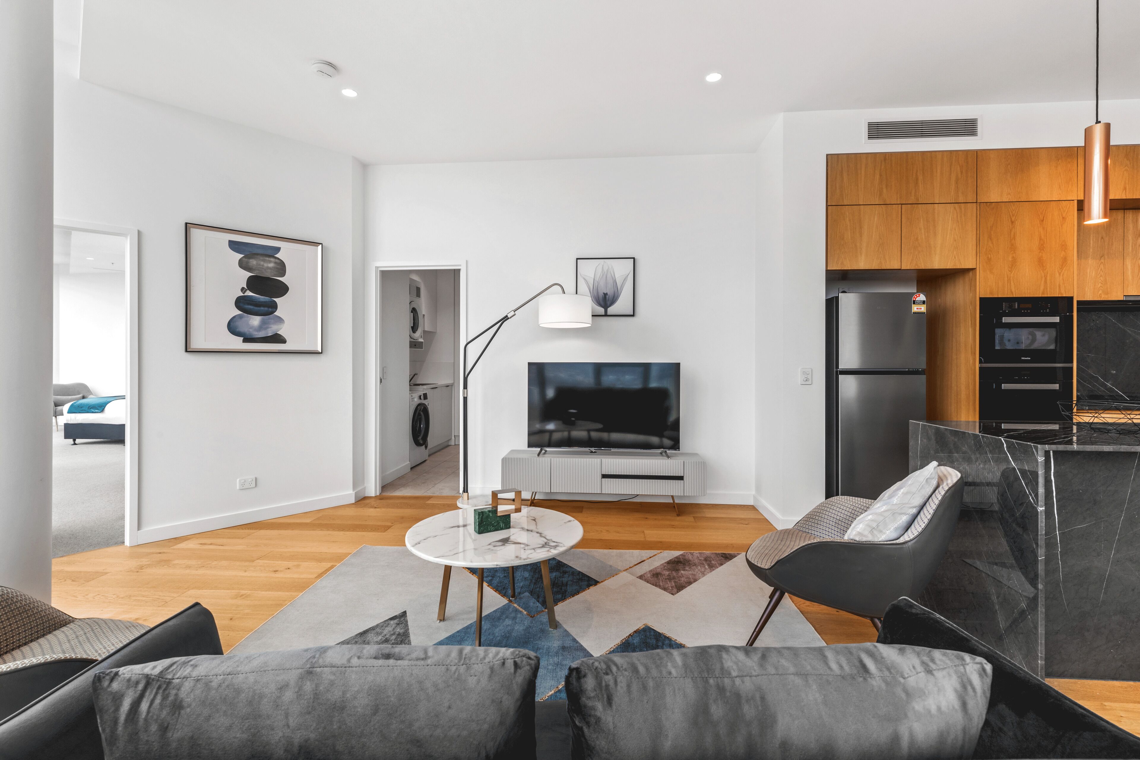 A modern living room with a sofa, a coffee table, and a TV. There is a kitchen area with wooden cabinets and a refrigerator.