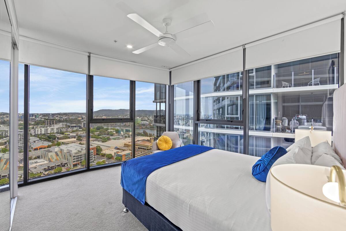 Habitación de hotel moderna con una cama grande, ventilador de techo y vistas a la ciudad a través de ventanales de piso a techo.
