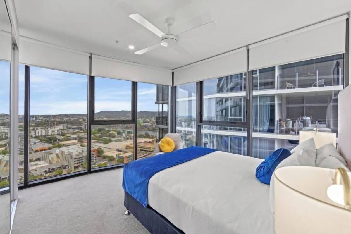 Un dormitorio moderno con una cama grande, un ventilador de techo y vistas a la ciudad a través de ventanales de piso a techo.