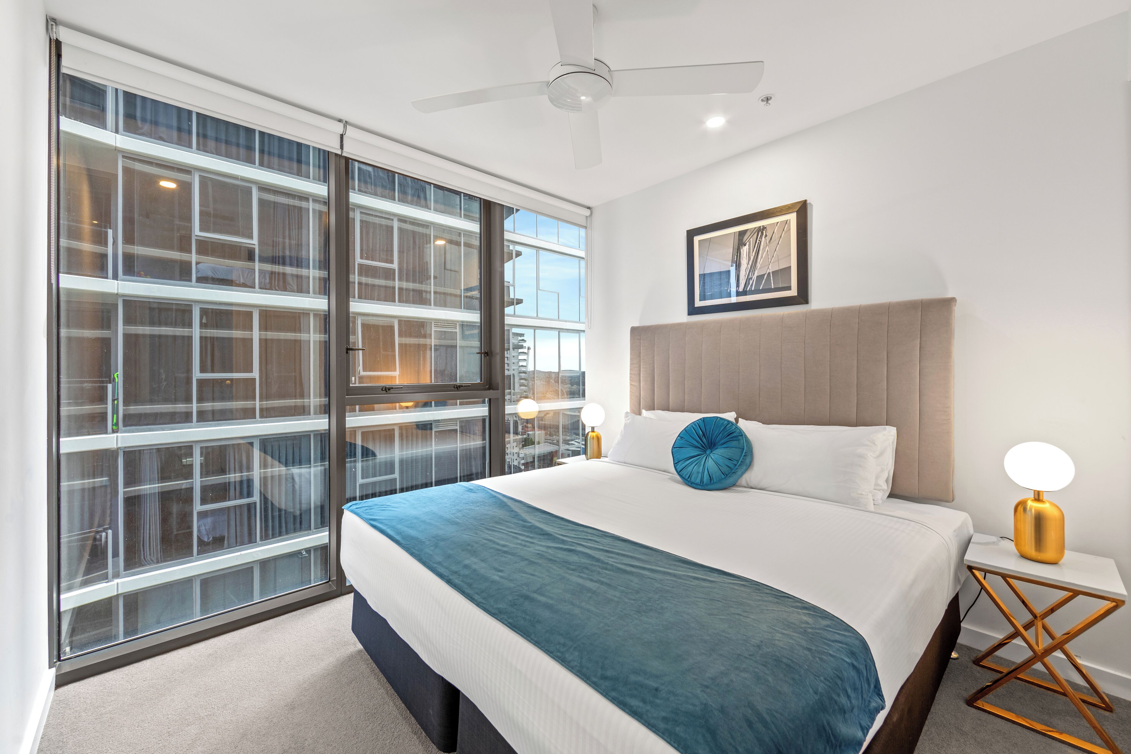 A modern bedroom with a large sliding glass door, a bed with a blue throw pillow, a bedside table with a lamp, and a ceiling fan.