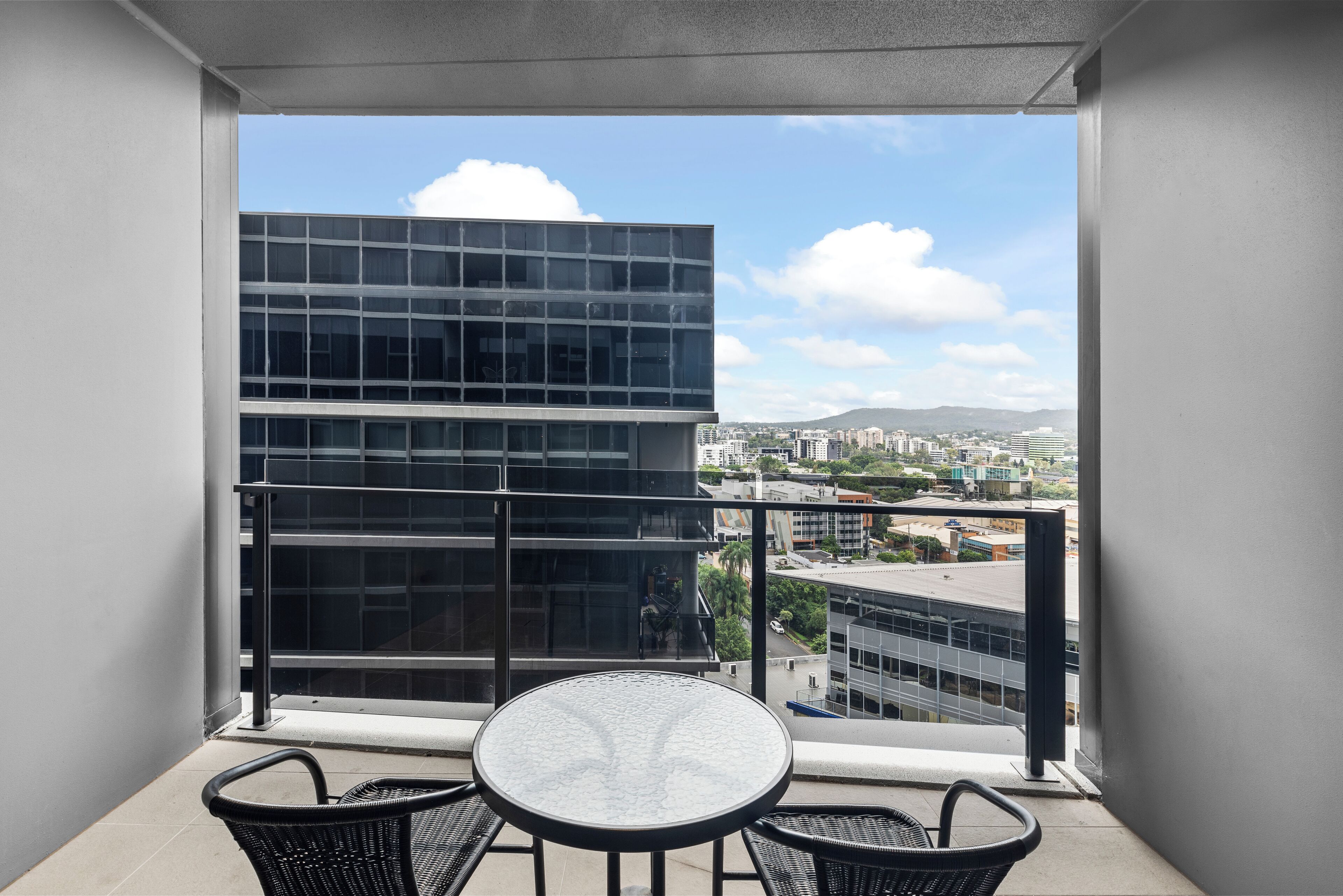 A balcony with a glass table and two chairs, overlooking a cityscape with modern buildings and a clear sky.