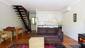 Una sala moderna con un sofá marrón, una mesa de comedor de madera, sillas de mimbre, una escalera, una cocina con armarios blancos y un hogar a leña.