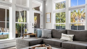 Un salon moderne avec un canapé d’angle gris, une table basse en osier et une porte vitrée donnant sur un balcon d’où l’on peut admirer des arbres et une maison.