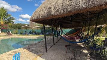 A pool area with a thatched roof structure, lounge chairs, and a hammock.