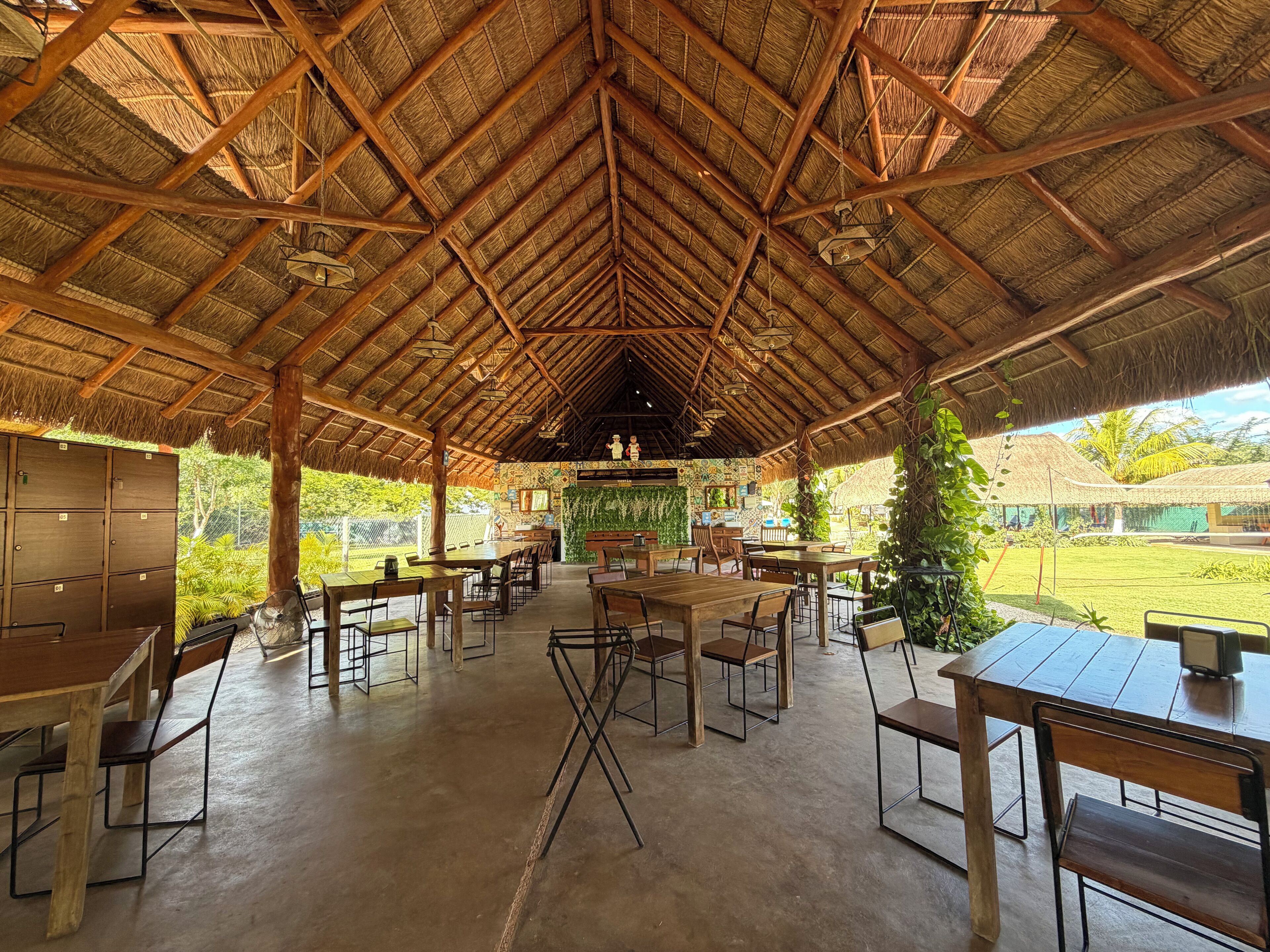 An open-air dining area with wooden tables and chairs, a thatched roof, and a view of greenery outside.