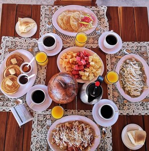 A breakfast spread on a wooden table with various dishes, drinks, and a decorative doily.