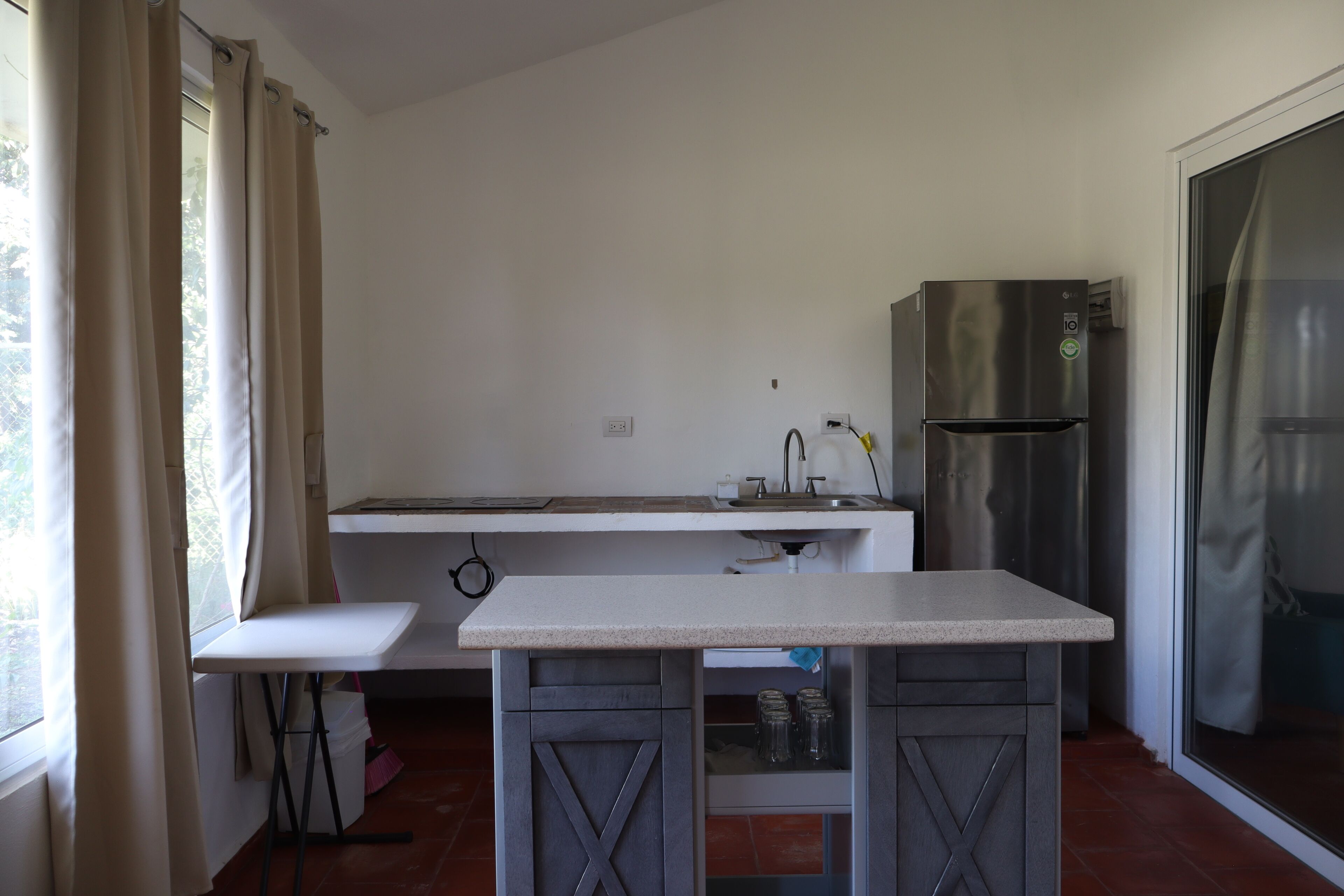 A compact kitchen with a white countertop, a stainless steel refrigerator, and a window with curtains.