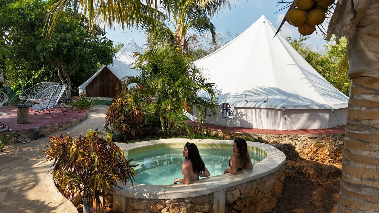 Un área de piscina con dos personas, rodeada de palmeras y una carpa blanca.