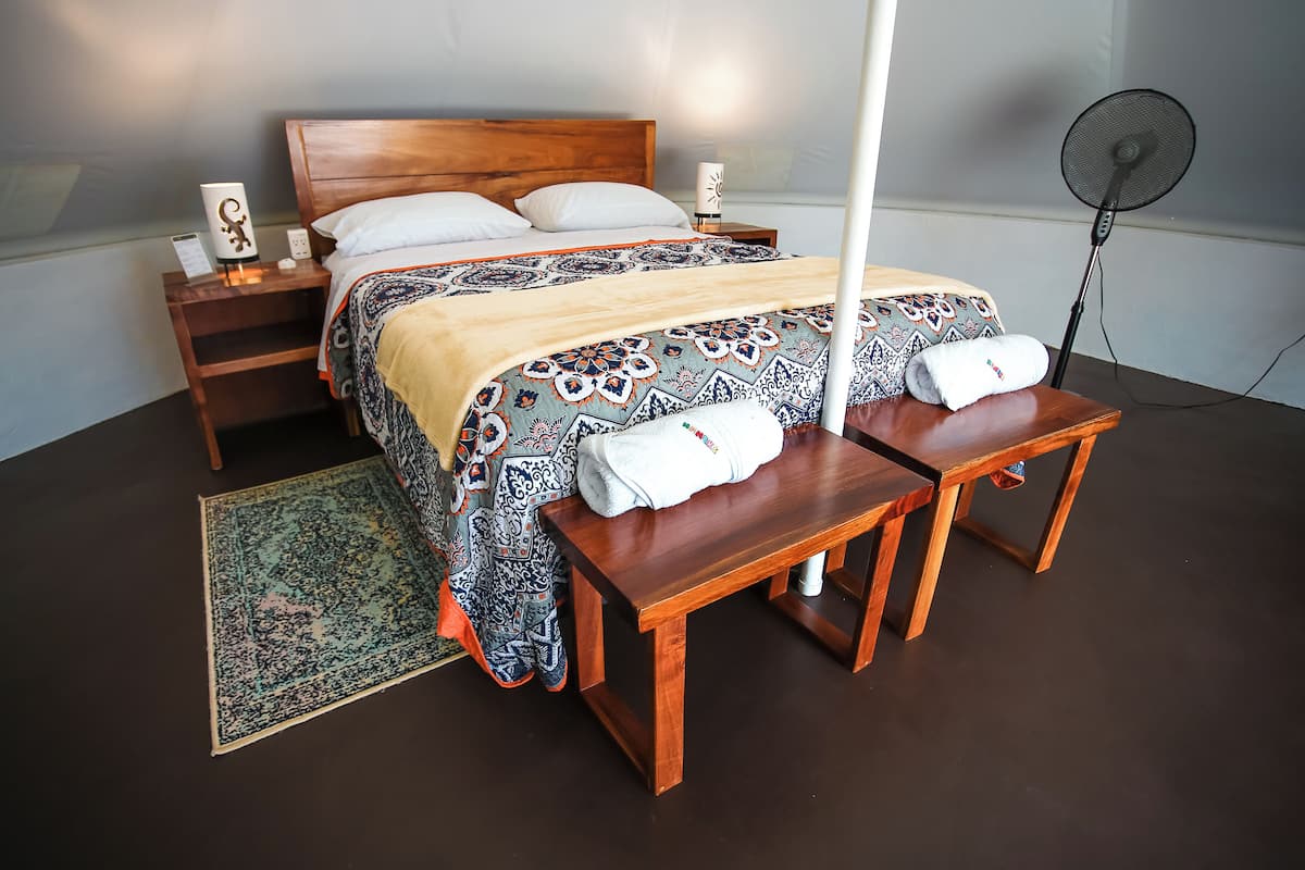 A bedroom with a wooden bed, bedside tables, a standing fan, and a patterned rug.