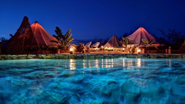 A nighttime resort with a swimming pool, thatched-roof structures, and well-lit pathways.