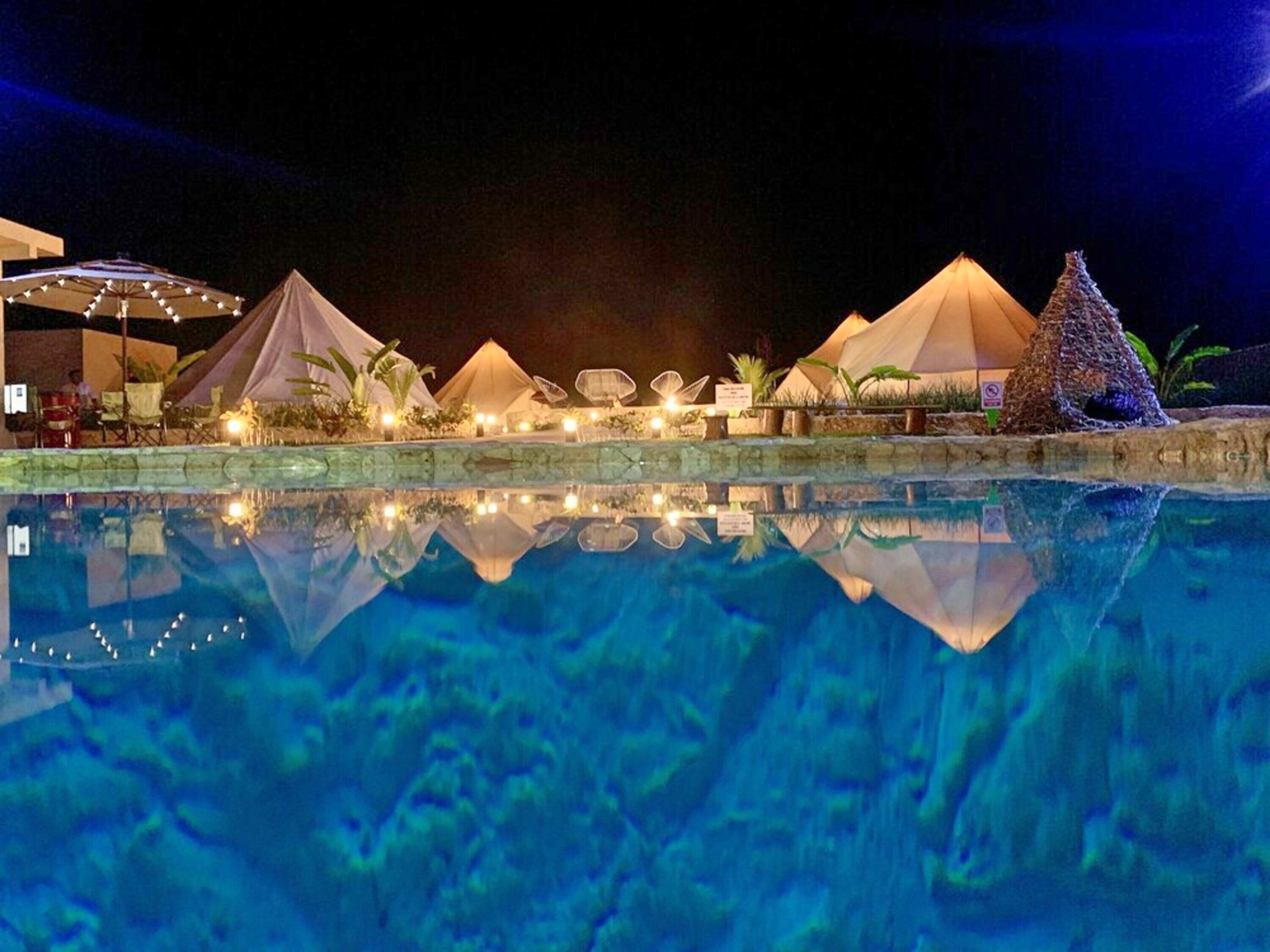 A nighttime view of a pool area with multiple tents and umbrellas, reflecting on the water.