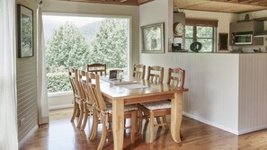 Un espace repas avec une table et des chaises en bois, une vue sur les arbres à travers de grandes fenêtres, et un coin cuisine avec des placards et des appareils électroménagers.