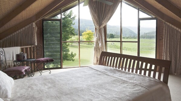 Une chambre avec une grande fenêtre, un lit en bois, une petite table avec une chaise, et une vue sur un paysage verdoyant.