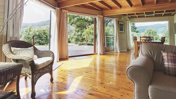 A spacious room with wooden floors, wicker furniture, and large windows offering a view of a mountainous landscape. - Cloud Nine Chalet - Luxury retreat in Upper River! (UPPER KANGAROO RIVER)