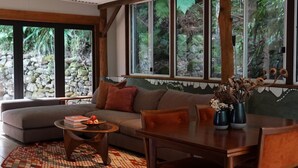 A cozy living room with a sectional sofa, a coffee table, and a dining table. Large windows offer a view of a stone wall and greenery outside. - Lyra - Kangaroo Valley (KANGAROO VALLEY)