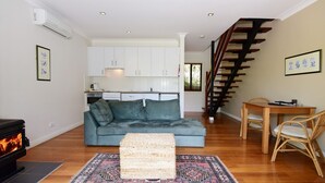 A modern living room with a sofa, a woven ottoman, a fireplace, a dining area with a table and chairs, a kitchen with white cabinets, and a wooden staircase. - Cloudsong Chalet 3 - Close to the village centre! (KANGAROO VALLEY)