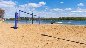 Un filet de volley-ball de plage sur lequel on peut lire le mot « BUDDY'S », une plage de sable et une étendue d'eau, avec des arbres et des bâtiments en arrière-plan.