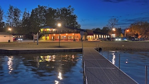 A waterfront area with a dock leading to a building with outdoor seating, surrounded by trees and a sandy area. - Lakefront • Hot Tub • Walk to Beach & Downtown (Lake Odessa)