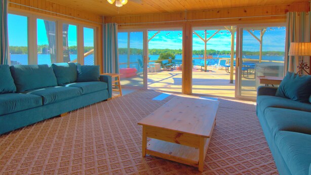 A spacious living room with a large sofa, a coffee table, and a view of a lakeside deck with outdoor seating. - Lakefront • Hot Tub • Walk to Beach & Downtown (Lake Odessa)